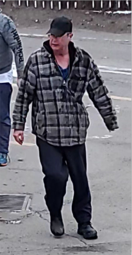 Un homme blanc , age moyen, vêtu d’une veste à carreaux brune et d’un pantalon foncé.