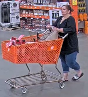 Femme de taille forte poussant un chariot de Home Depot à la sortie de Home Depot. Son chariot est maintenant rempli d’outils. Elle a de longs cheveux bruns qui ont été placés en chignon et porte un jean bleu, une chemise noire à manches longues et des sandales noires à rayures blanches.