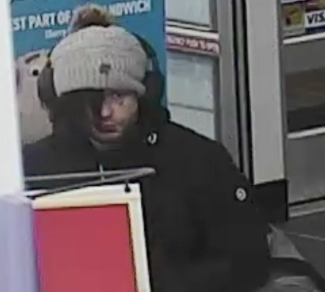 Le suspect fait face à la caméra; il porte une tuque surmontée d’un casque d’écoute. Ses cheveux dépassent de la tuque et lui recouvrent un œil. Il porte également un manteau noir. 